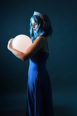 Mavi saçlı, göz kamaştırıcı fantezi balo elbisesi giyen, kristal taç takan, sanatsal duruşta parıldayan lamba taşıyan güzel bir kadın modelin portresi. Karanlık stüdyo arka planında izole.