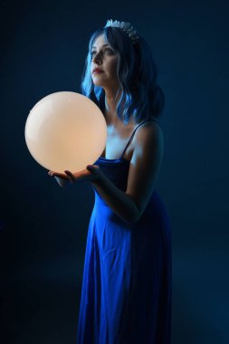 Mavi saçlı, göz kamaştırıcı fantezi balo elbisesi giyen, kristal taç takan, sanatsal duruşta parıldayan lamba taşıyan güzel bir kadın modelin portresi. Karanlık stüdyo arka planında izole.