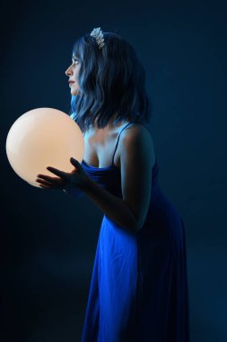 Mavi saçlı, göz kamaştırıcı fantezi balo elbisesi giyen, kristal taç takan, sanatsal duruşta parıldayan lamba taşıyan güzel bir kadın modelin portresi. Karanlık stüdyo arka planında izole.