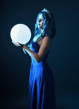 Mavi saçlı, göz kamaştırıcı fantezi balo elbisesi giyen, kristal taç takan, sanatsal duruşta parıldayan lamba taşıyan güzel bir kadın modelin portresi. Karanlık stüdyo arka planında izole.