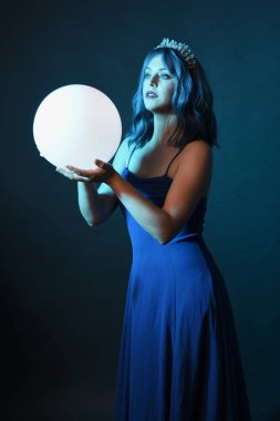Mavi saçlı, göz kamaştırıcı fantezi balo elbisesi giyen, kristal taç takan, sanatsal duruşta parıldayan lamba taşıyan güzel bir kadın modelin portresi. Karanlık stüdyo arka planında izole.
