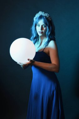 Mavi saçlı, göz kamaştırıcı fantezi balo elbisesi giyen, kristal taç takan, sanatsal duruşta parıldayan lamba taşıyan güzel bir kadın modelin portresi. Karanlık stüdyo arka planında izole.