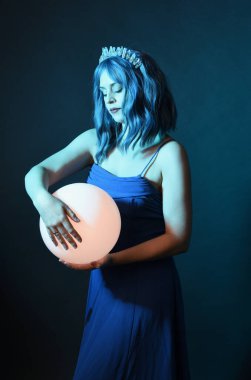 Mavi saçlı, göz kamaştırıcı fantezi balo elbisesi giyen, kristal taç takan, sanatsal duruşta parıldayan lamba taşıyan güzel bir kadın modelin portresi. Karanlık stüdyo arka planında izole.