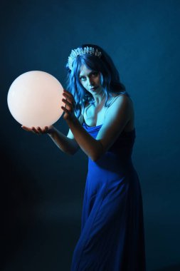 Mavi saçlı, göz kamaştırıcı fantezi balo elbisesi giyen, kristal taç takan, sanatsal duruşta parıldayan lamba taşıyan güzel bir kadın modelin portresi. Karanlık stüdyo arka planında izole.