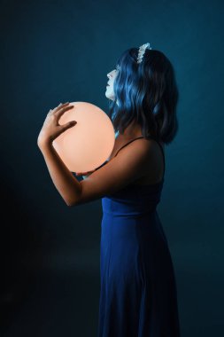 Mavi saçlı, göz kamaştırıcı fantezi balo elbisesi giyen, kristal taç takan, sanatsal duruşta parıldayan lamba taşıyan güzel bir kadın modelin portresi. Karanlık stüdyo arka planında izole.