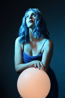Mavi saçlı, göz kamaştırıcı fantezi balo elbisesi giyen, kristal taç takan, sanatsal duruşta parıldayan lamba taşıyan güzel bir kadın modelin portresi. Karanlık stüdyo arka planında izole.