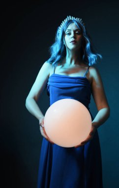 Mavi saçlı, göz kamaştırıcı fantezi balo elbisesi giyen, kristal taç takan, sanatsal duruşta parıldayan lamba taşıyan güzel bir kadın modelin portresi. Karanlık stüdyo arka planında izole.