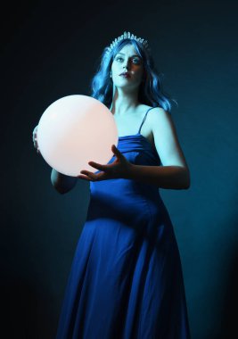 Mavi saçlı, göz kamaştırıcı fantezi balo elbisesi giyen, kristal taç takan, sanatsal duruşta parıldayan lamba taşıyan güzel bir kadın modelin portresi. Karanlık stüdyo arka planında izole.