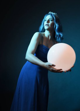Mavi saçlı, göz kamaştırıcı fantezi balo elbisesi giyen, kristal taç takan, sanatsal duruşta parıldayan lamba taşıyan güzel bir kadın modelin portresi. Karanlık stüdyo arka planında izole.