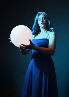Mavi saçlı, göz kamaştırıcı fantezi balo elbisesi giyen, kristal taç takan, sanatsal duruşta parıldayan lamba taşıyan güzel bir kadın modelin portresi. Karanlık stüdyo arka planında izole.