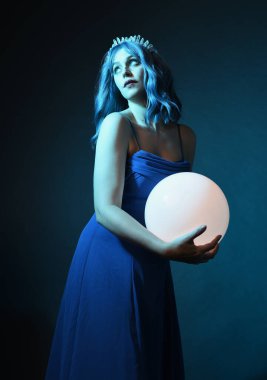 Mavi saçlı, göz kamaştırıcı fantezi balo elbisesi giyen, kristal taç takan, sanatsal duruşta parıldayan lamba taşıyan güzel bir kadın modelin portresi. Karanlık stüdyo arka planında izole.