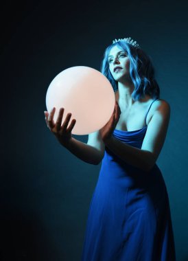 Mavi saçlı, göz kamaştırıcı fantezi balo elbisesi giyen, kristal taç takan, sanatsal duruşta parıldayan lamba taşıyan güzel bir kadın modelin portresi. Karanlık stüdyo arka planında izole.