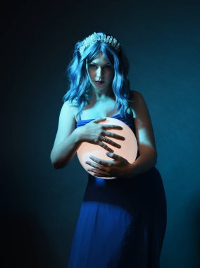 Mavi saçlı, göz kamaştırıcı fantezi balo elbisesi giyen, kristal taç takan, sanatsal duruşta parıldayan lamba taşıyan güzel bir kadın modelin portresi. Karanlık stüdyo arka planında izole.