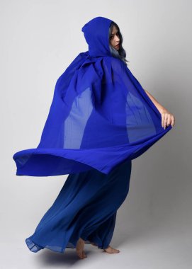 Güzel, genç, zarif, mavi fantezi balo elbisesi ve kapüşonlu bir pelerinin tam boy portresi. Duruşunu, arkasını dönüp uzaklaşmasını. Whit stüdyosunun arka planında izole..