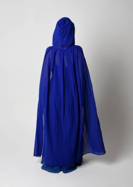 Güzel, genç, zarif, mavi fantezi balo elbisesi ve kapüşonlu bir pelerinin tam boy portresi. Duruşunu, arkasını dönüp uzaklaşmasını. Whit stüdyosunun arka planında izole..