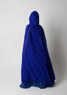 Güzel, genç, zarif, mavi fantezi balo elbisesi ve kapüşonlu bir pelerinin tam boy portresi. Duruşunu, arkasını dönüp uzaklaşmasını. Whit stüdyosunun arka planında izole..