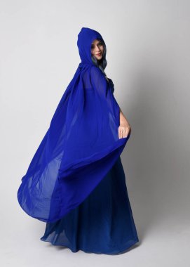 Full length portrait of beautiful young female model elegant blue fantasy  ball gown and flowing cape with hood. Standing pose, walking away facing backwards. Isolated on whit studio background. clipart