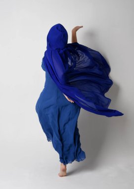 Güzel, genç, zarif, mavi fantezi balo elbisesi ve kapüşonlu bir pelerinin tam boy portresi. Duruşunu, arkasını dönüp uzaklaşmasını. Whit stüdyosunun arka planında izole..