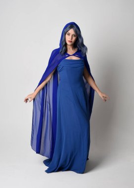 Zarif bir fantezi mavi balo elbisesi giyen ve kapüşonlu dalgalı bir pelerin giyen güzel bir kadın modelin tam boy portresi. Duruş, hareketli kollar uzanıyor. Beyaz stüdyo arka planında izole edilmiş..