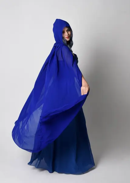 stock image Full length portrait of beautiful young female model elegant blue fantasy  ball gown and flowing cape with hood. Standing pose, walking away facing backwards. Isolated on whit studio background.