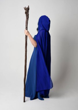 Full length portrait of beautiful female model wearing elegant fantasy blue ball gown, flowing cape with hood.Standing pose walking away, holding a wooden wizard staff. Isolated on white studio background. clipart