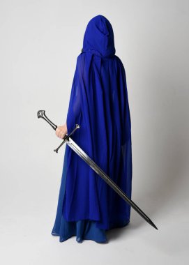 Zarif fantezi mavi balo elbisesi giyen, kapüşonlu pelerin giyen güzel bir kadın modelin tam boy portresi. Beyaz stüdyo arka planında izole edilmiş bir kılıçla uzaklaşıyor..