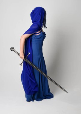 Zarif fantezi mavi balo elbisesi giyen, kapüşonlu pelerin giyen güzel bir kadın modelin tam boy portresi. Beyaz stüdyo arka planında izole edilmiş bir kılıçla uzaklaşıyor..