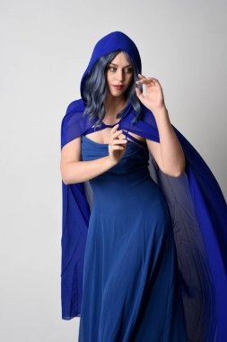 Zarif fantezi mavi balo elbisesi giyen, kapüşonlu pelerinli güzel bir kadın modelin portresi. El kol hareketleri. Beyaz stüdyo arka planında izole edilmiş..