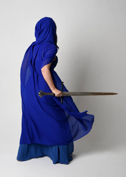 Full length portrait of beautiful female model wearing elegant fantasy blue ball gown, flowing cape with hood.Standing pose walking away, holding a sword weapon Isolated on white studio background.