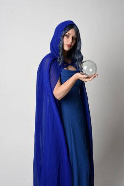 Zarif fantezi mavi balo elbisesi giyen kapüşonlu pelerinli güzel bir kadın modelin portresi. Kristal cam bir kahin küresi tutuyorum. Beyaz stüdyo arka planında izole edilmiş..