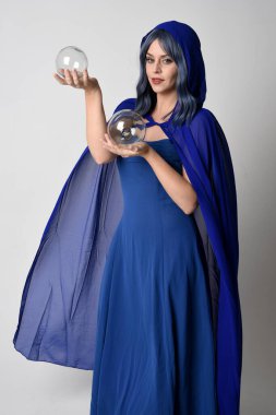 Zarif fantezi mavi balo elbisesi giyen kapüşonlu pelerinli güzel bir kadın modelin portresi. Kristal cam bir kahin küresi tutuyorum. Beyaz stüdyo arka planında izole edilmiş..