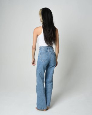 Esmer kadın modelin gündelik giysiler, beyaz atlet gömlek ve kot pantolon giymiş tam boy portresi. Kameradan uzaklaşarak poz veriyor. Beyaz stüdyo arka planında izole edilmiş..