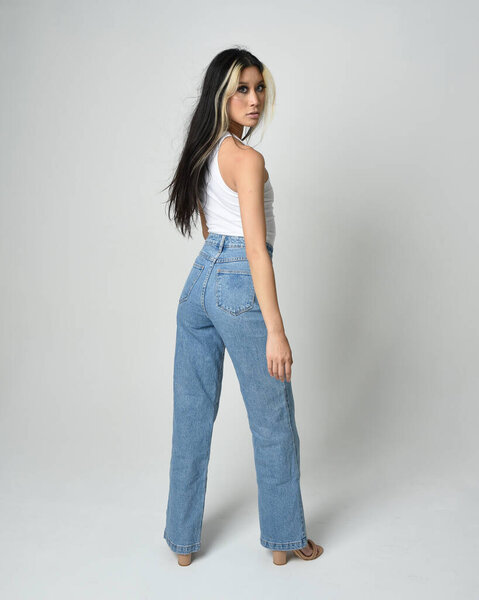 Полноразмерный портрет брюнетки-модели в повседневной одежде, белой одноразовой рубашке, джинсовых брюках. Стоячая поза уходит от камеры. Изолированный на белом фоне студии.