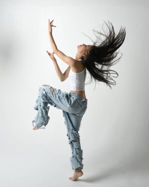 Esmer kadın Asyalı modelin günlük giysiler, beyaz atlet gömlek ve kot pantolon giymiş tam boy portresi. Enerjik zıplama, dans pozisyonu. Beyaz stüdyo arka planında izole edilmiş..