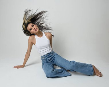 Esmer kadın Asyalı modelin günlük giysiler, beyaz atlet gömlek ve kot pantolon giymiş tam boy portresi. Oturma pozisyonu, perspektif için yüksek kamera açısı. Beyaz stüdyo arka planında izole edilmiş..