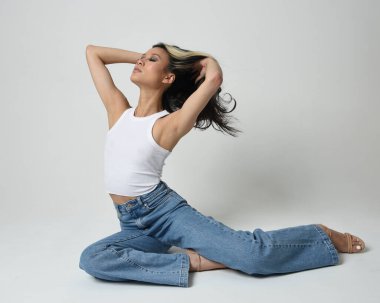 Esmer kadın Asyalı modelin günlük giysiler, beyaz atlet gömlek ve kot pantolon giymiş tam boy portresi. Oturma pozisyonu, perspektif için yüksek kamera açısı. Beyaz stüdyo arka planında izole edilmiş..