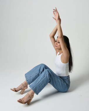 Esmer kadın Asyalı modelin günlük giysiler, beyaz atlet gömlek ve kot pantolon giymiş tam boy portresi. Oturma pozisyonu, perspektif için yüksek kamera açısı. Beyaz stüdyo arka planında izole edilmiş..