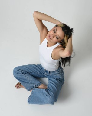 Esmer kadın Asyalı modelin günlük giysiler, beyaz atlet gömlek ve kot pantolon giymiş tam boy portresi. Oturma pozisyonu, perspektif için yüksek kamera açısı. Beyaz stüdyo arka planında izole edilmiş..