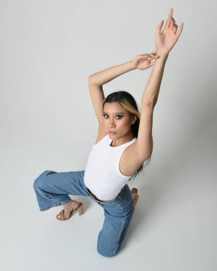 Esmer kadın Asyalı modelin günlük giysiler, beyaz atlet gömlek ve kot pantolon giymiş tam boy portresi. Oturma pozisyonu, perspektif için yüksek kamera açısı. Beyaz stüdyo arka planında izole edilmiş..