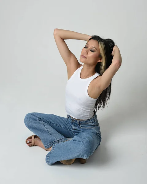 stock image Full length portrait of brunette female asian model wearing casual clothes, white singlet shirt, denim jean pants. Sitting pose, high camera angle for perspective. Isolated on white studio background.