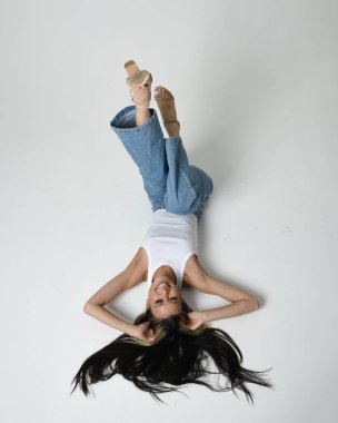 Esmer kadın Asyalı modelin günlük giysiler, beyaz atlet gömlek ve kot pantolon giymiş tam boy portresi. Oturma pozisyonu, yan yatmış, bacakları havada. perspektif için yüksek kamera açısı. Beyaz stüdyo arka planında izole edilmiş..