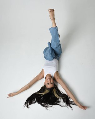 Esmer kadın Asyalı modelin günlük giysiler, beyaz atlet gömlek ve kot pantolon giymiş tam boy portresi. Oturma pozisyonu, yan yatmış, bacakları havada. perspektif için yüksek kamera açısı. Beyaz stüdyo arka planında izole edilmiş..