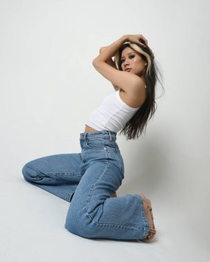 Esmer kadın Asyalı modelin günlük giysiler, beyaz atlet gömlek ve kot pantolon giymiş tam boy portresi. Rahat bir poz. Stüdyo arka planında izole edilmiş.