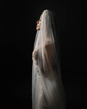 Beyaz gelinlik giymiş güzel bir mankenin yakın portresi ve kefen gibi akan bir duvak. Huysuz sinematik ışıklandırma, karanlık bir stüdyonun arka planında izole edilmiş..