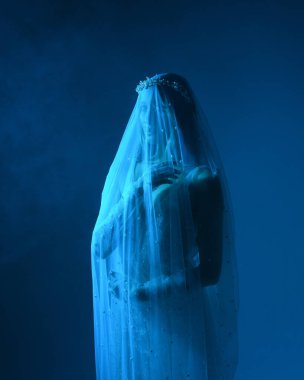 Beyaz gelinlik giymiş güzel bir mankenin yakın portresi ve kefen gibi akan bir duvak. Huysuz sinematik ışıklandırma, karanlık bir stüdyonun arka planında izole edilmiş..