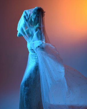 Beyaz gelinlik giymiş güzel bir mankenin yakın portresi ve kefen gibi akan bir duvak. Huysuz sinematik ışıklandırma, karanlık bir stüdyonun arka planında izole edilmiş..