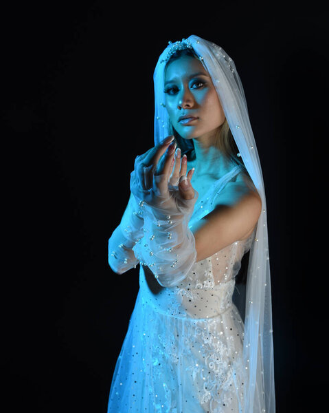 Close up portrait of beautiful model wearing white wedding gown and ghostly flowing veil. Posing with gestural hands reaching out beckoning.  Moody cinematic lighting, isolated on a dark studio background.