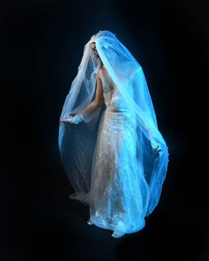 Beyaz gelinlik giymiş güzel bir mankenin uzun bir portresi ve kefen gibi akan bir duvak. Huysuz sinematik ışıklandırma, karanlık bir stüdyonun arka planında izole edilmiş..