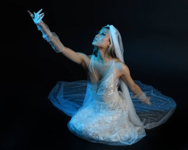 Beyaz gelinlik giymiş güzel bir mankenin uzun bir portresi ve kefen gibi akan bir duvak. Huysuz sinematik ışıklandırma, oturma pozu, karanlık bir stüdyo arka planında izole edilmiş..