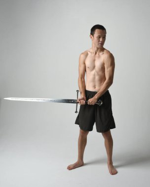 Yakışıklı Asyalı erkek modelin tam boy portresi, spor şortu ve gömleksiz. Beyaz stüdyo arka planında izole edilmiş, savaşçı eğitimi sırasında kılıçlarını tutuyor..